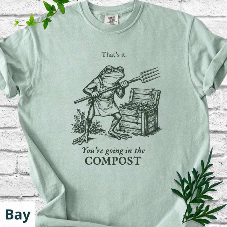 May include: A light green t-shirt featuring a frog illustration holding a pitchfork, standing near a wooden compost bin. The text above the frog reads "That's it." and below it, "You're going in the COMPOST."