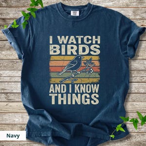 May include: Navy blue t-shirt with the text "I WATCH BIRDS AND I KNOW THINGS." The design features a bird perched on a branch with leaves and berries, set against a retro-style striped background in orange, yellow, and brown.