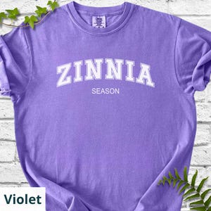 May include: A violet t-shirt featuring the word "ZINNIA" in white, arched lettering, with "SEASON" underneath. The long-sleeved shirt is a solid colour and has a crew neck.