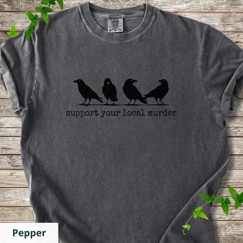 May include: A charcoal gray short-sleeve t-shirt featuring a black graphic of four crows standing in a row. Below the birds, the text reads "support your local murder" in a typewriter-style font. The shirt has a crew neckline and a washed finish.