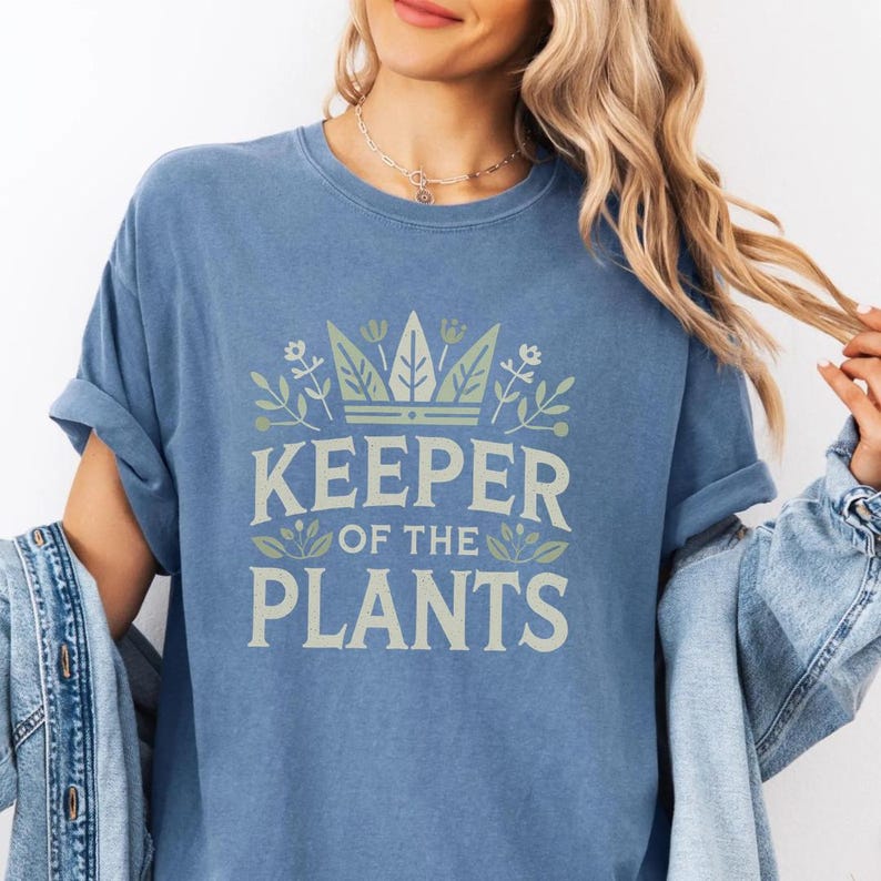 May include: A blue t-shirt with the text "KEEPER OF THE PLANTS" in a light green font. The design includes a crown and floral elements. The shirt has short sleeves and a relaxed fit.