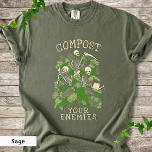 Peut inclure: T-shirt vert sauge avec un motif de trois squelettes entour&eacute;s de feuillage vert. Le texte "COMPOST YOUR ENEMIES" est imprim&eacute; au-dessus et en dessous des squelettes. Le t-shirt a un col rond et des manches courtes.