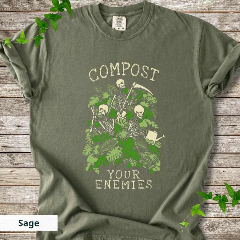 Peut inclure: T-shirt vert sauge avec un motif de trois squelettes entour&eacute;s de feuillage vert. Le texte "COMPOST YOUR ENEMIES" est imprim&eacute; au-dessus et en dessous des squelettes. Le t-shirt a un col rond et des manches courtes.