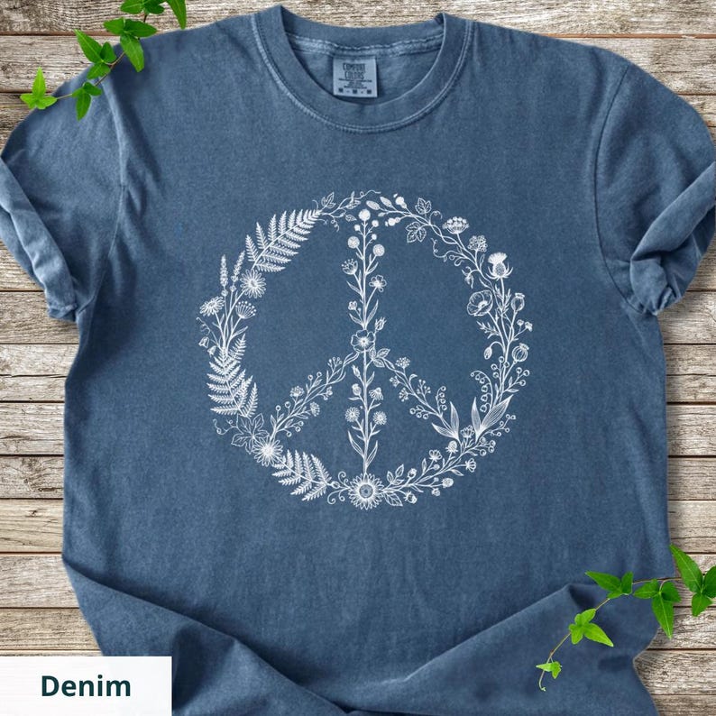 May include: A blue short-sleeved t-shirt featuring a white graphic print of a peace sign composed of various botanical elements, including ferns, wildflowers, and leafy stems. The shirt has a crew neckline and a label reading Comfort Colors.