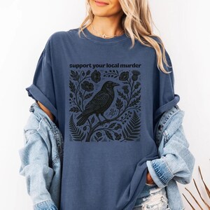 May include: A slate blue short-sleeve t-shirt featuring a black graphic print of a crow perched on a branch surrounded by botanical elements like ferns and flowers. The text "support your local murder" is printed in black above the illustration.