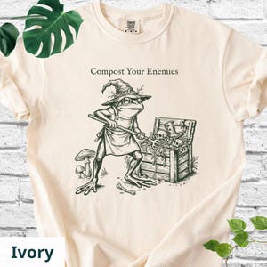 May include: Ivory-colored t-shirt featuring a whimsical illustration of a frog wearing a witch's hat and apron, stirring a chest of bones. The text "Compost Your Enemies" is printed above the frog. The shirt has a crew neck and short sleeves.