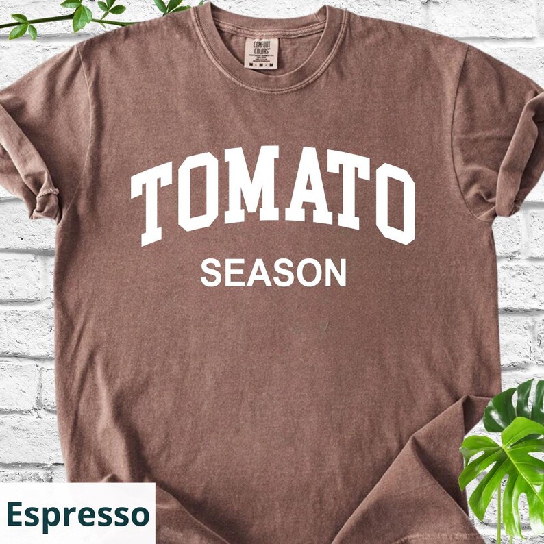 K&ouml;nnte beinhalten: Espressofarbenes T-Shirt mit den Worten "TOMATO SEASON" in wei&szlig;en Blockbuchstaben. Das Shirt hat einen Rundhalsausschnitt und kurze &Auml;rmel. Das Shirt ist aus einem weichen, bequemen Material.