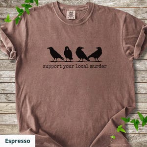 May include: A brown short-sleeve t-shirt featuring a black graphic of four crows standing in a row. Below the birds, the text reads "support your local murder" in a typewriter-style font. The shirt has a crew neckline and a visible brand label.