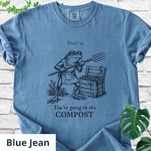 May include: Blue jean colored t-shirt featuring a frog with a pitchfork standing in front of a compost bin. The shirt has the text "That's it." above the frog and "You're going in the COMPOST" below.