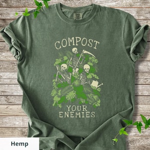 Peut inclure: T-shirt vert olive avec l'inscription "COMPOST YOUR ENEMIES" en police vieillie. Le motif repr&eacute;sente des squelettes parmi un feuillage vert, l'un tenant une faux et l'autre un arrosoir. Le mot "Hemp" est en bas.