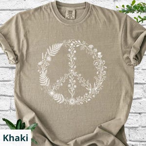 May include: A khaki-colored short-sleeve t-shirt featuring a white graphic print of a peace sign composed of various botanical elements, including ferns, wildflowers, and stems. The shirt has a crew neckline and a visible brand label at the neck.