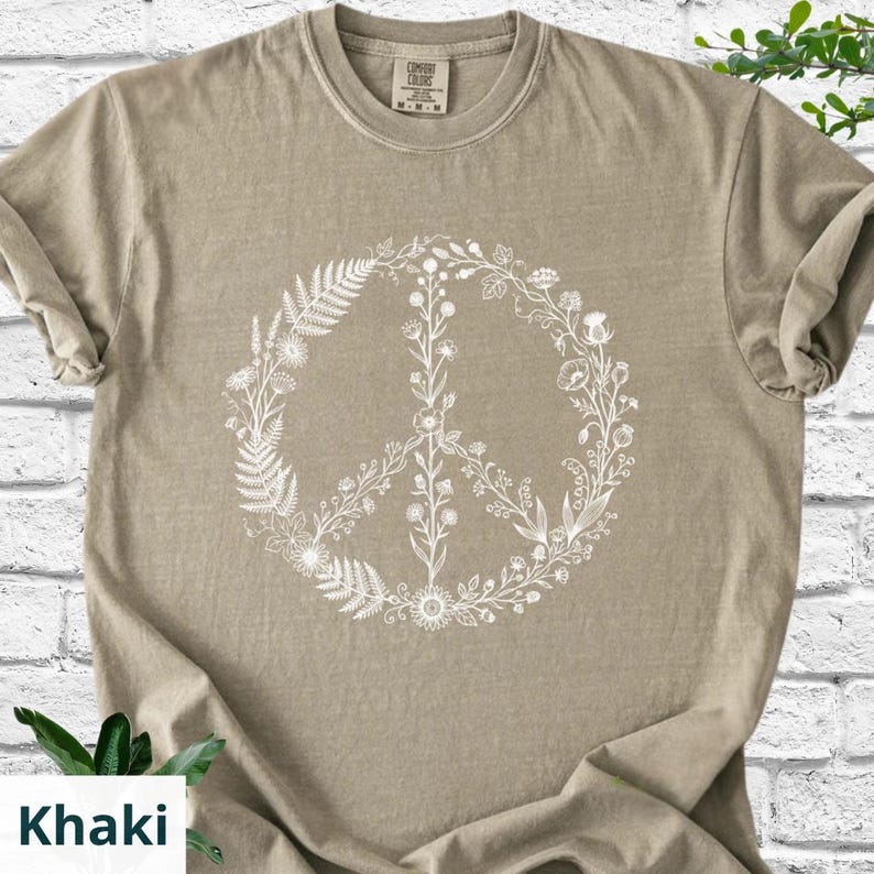 May include: A khaki-colored short-sleeve t-shirt featuring a white graphic print of a peace sign composed of various botanical elements, including ferns, wildflowers, and stems. The shirt has a crew neckline and a visible brand label at the neck.
