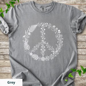 May include: A grey short-sleeved t-shirt featuring a large white graphic print of a peace sign composed of various botanical elements including ferns, wildflowers, and stems. The shirt has a crew neckline and a visible brand tag at the collar.