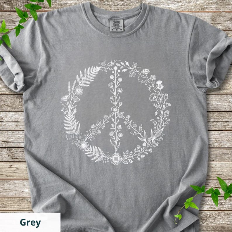 May include: A grey short-sleeved t-shirt featuring a large white graphic print of a peace sign composed of various botanical elements including ferns, wildflowers, and stems. The shirt has a crew neckline and a visible brand tag at the collar.