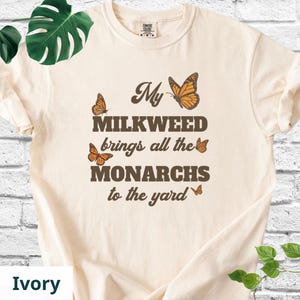 May include: Ivory t-shirt with the text "My MILKWEED brings all the MONARCHS to the yard" in brown lettering. The shirt features illustrations of orange butterflies. The shirt is a Comfort Colors brand.