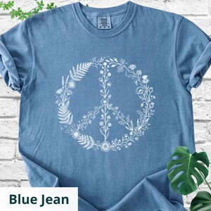 May include: A blue jean colored short-sleeved t-shirt featuring a white graphic print of a peace sign composed of various botanical elements, including ferns, wildflowers, and leafy stems arranged in a circular shape on the front of the garment.