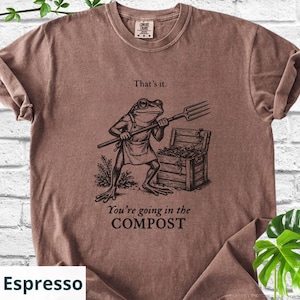 May include: Espresso-colored t-shirt featuring a frog character holding a pitchfork, standing near a wooden compost bin. The shirt has the phrases "That's it." and "You're going in the COMPOST" printed on it.