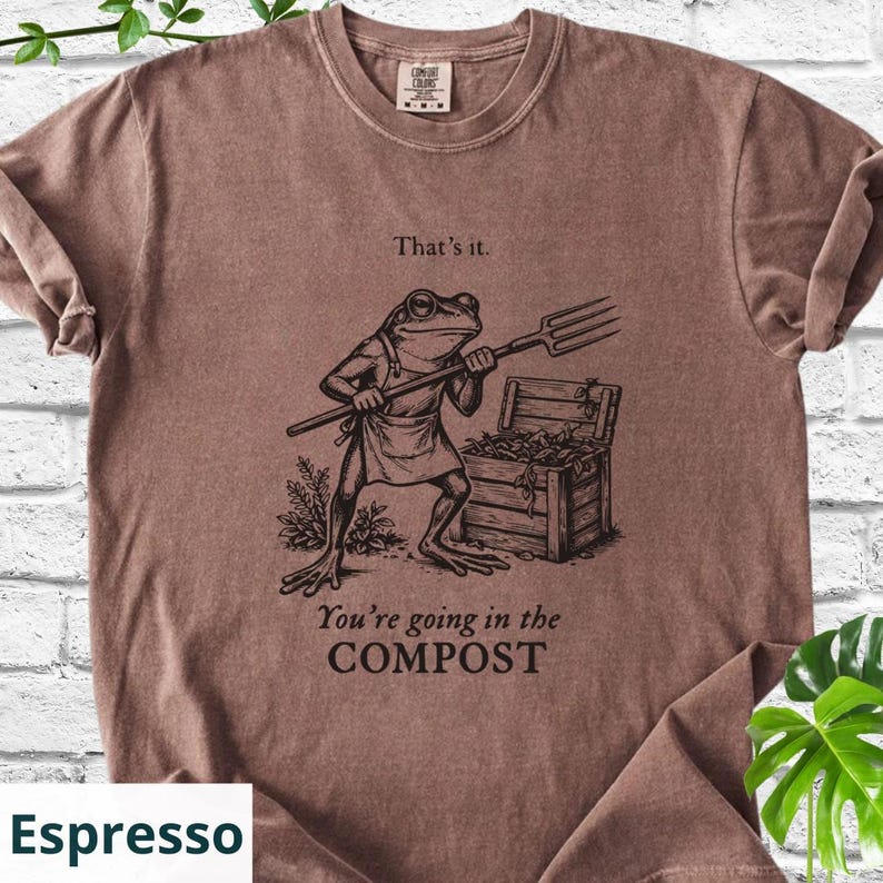 May include: Espresso-colored t-shirt featuring a frog character holding a pitchfork, standing near a wooden compost bin. The shirt has the phrases "That's it." and "You're going in the COMPOST" printed on it.