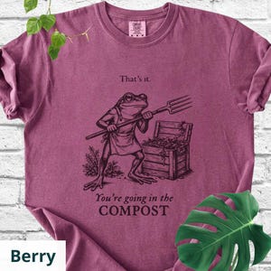 May include: Berry-colored t-shirt featuring a humorous illustration of a frog holding a pitchfork. The frog stands in front of a compost bin, with the text "That's it." and "You're going in the COMPOST" printed below the frog.