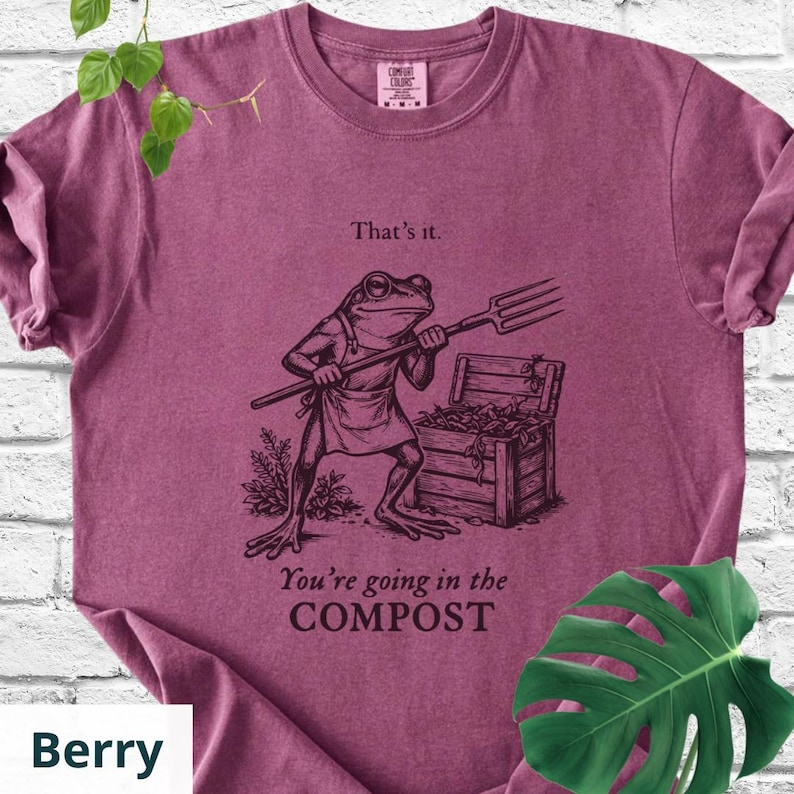 May include: Berry-colored t-shirt featuring a humorous illustration of a frog holding a pitchfork. The frog stands in front of a compost bin, with the text "That's it." and "You're going in the COMPOST" printed below the frog.