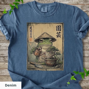 May include: Denim blue t-shirt featuring a cartoon frog wearing a conical hat, watering a bonsai tree. The frog is depicted with a watering can and a small shovel. Japanese text is visible on the design. The shirt is made of a soft material.