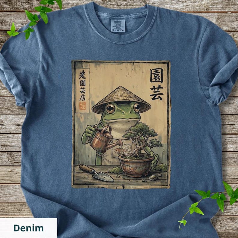 May include: Denim blue t-shirt featuring a cartoon frog wearing a conical hat, watering a bonsai tree. The frog is depicted with a watering can and a small shovel. Japanese text is visible on the design. The shirt is made of a soft material.