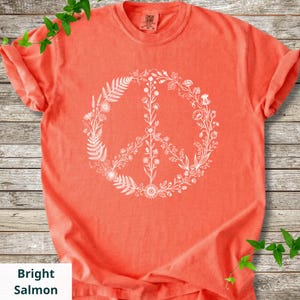 May include: A bright salmon-colored short-sleeve t-shirt featuring a large, white, circular peace sign graphic composed of various botanical illustrations, including ferns, wildflowers, and stems, centered on the front of the garment.