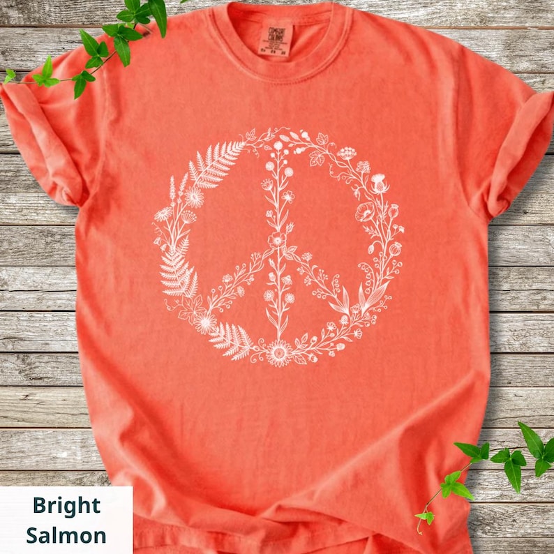 May include: A bright salmon-colored short-sleeve t-shirt featuring a large, white, circular peace sign graphic composed of various botanical illustrations, including ferns, wildflowers, and stems, centered on the front of the garment.