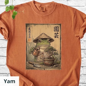 May include: Orange t-shirt featuring a detailed illustration of a frog wearing a conical hat, watering a bonsai tree. The artwork includes Japanese text and a small shovel. The shirt is made of a soft material.