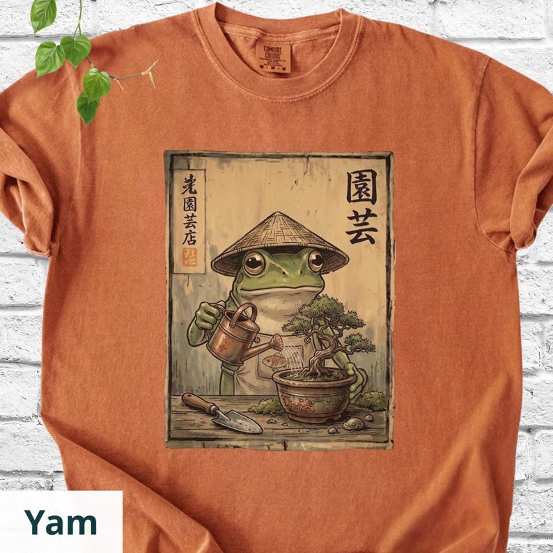 May include: Orange t-shirt featuring a detailed illustration of a frog wearing a conical hat, watering a bonsai tree. The artwork includes Japanese text and a small shovel. The shirt is made of a soft material.
