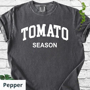 K&ouml;nnte beinhalten: Dunkelgraues T-Shirt mit dem wei&szlig;en Schriftzug "TOMATO SEASON" in Gro&szlig;buchstaben. Das Shirt hat einen Rundhalsausschnitt und kurze &Auml;rmel. Der Stoff scheint vorgeschrumpfte Baumwolle zu sein.