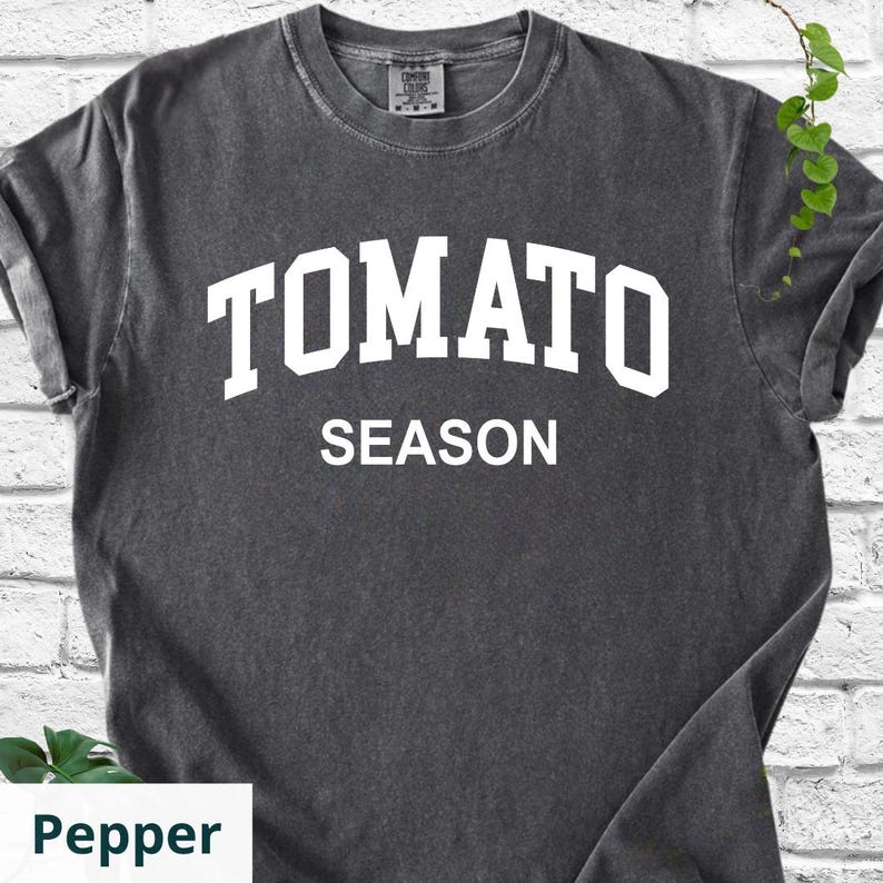 K&ouml;nnte beinhalten: Dunkelgraues T-Shirt mit dem wei&szlig;en Schriftzug "TOMATO SEASON" in Gro&szlig;buchstaben. Das Shirt hat einen Rundhalsausschnitt und kurze &Auml;rmel. Der Stoff scheint vorgeschrumpfte Baumwolle zu sein.
