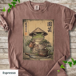 May include: Espresso-colored t-shirt featuring a cartoon frog wearing a conical hat, watering a bonsai tree. The frog is depicted with a watering can and a small shovel. Japanese text is visible on the design.