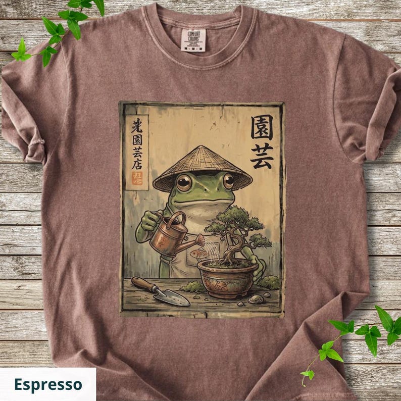 May include: Espresso-colored t-shirt featuring a cartoon frog wearing a conical hat, watering a bonsai tree. The frog is depicted with a watering can and a small shovel. Japanese text is visible on the design.