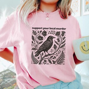 May include: A light pink short-sleeve t-shirt featuring a black graphic print of a crow perched on a branch surrounded by flowers and foliage. The text "support your local murder" is printed in black above the detailed botanical illustration.