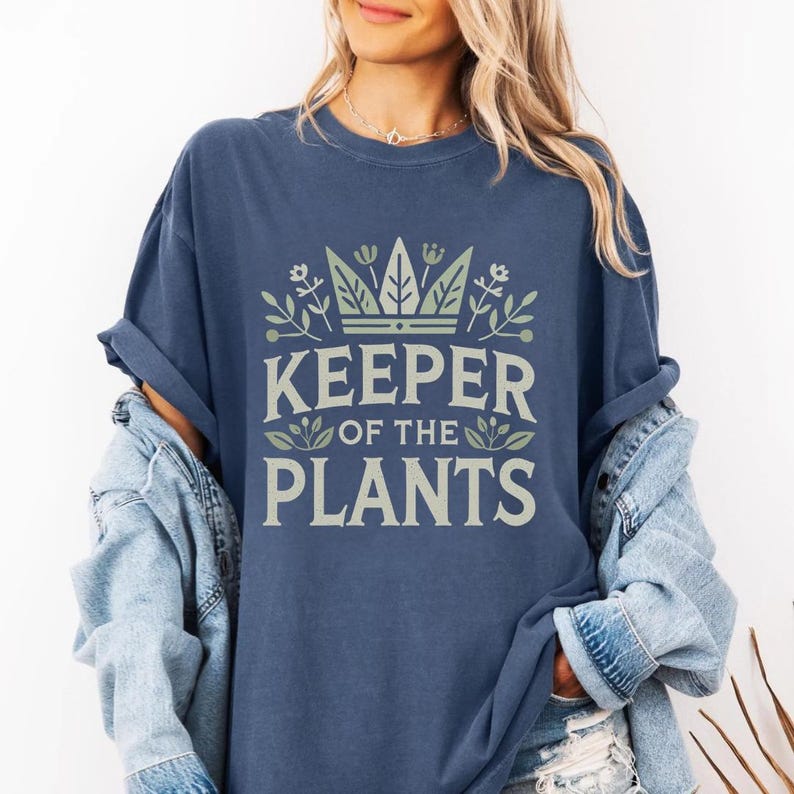 May include: A navy blue t-shirt with the text "KEEPER OF THE PLANTS" in a light green and white design. The design includes a crown and floral elements. The shirt is part of a casual clothing collection.