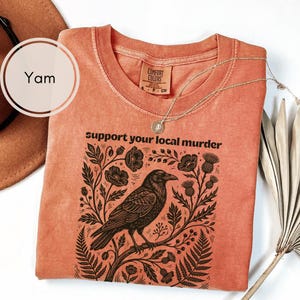 May include: A yam-colored crew neck t-shirt featuring a black graphic print of a crow perched on a branch surrounded by flowers and foliage. The text "support your local murder" is printed above the graphic in a black sans-serif font.