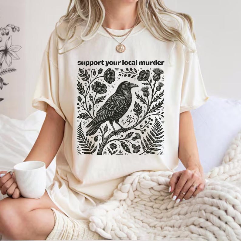 May include: A cream-colored t-shirt featuring a black graphic print of a crow perched on a branch surrounded by flowers and foliage. The text "support your local murder" is printed in black above the illustration in a simple sans-serif font.