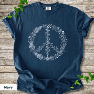 May include: A navy blue short-sleeve t-shirt featuring a white graphic print of a peace sign composed of various botanical elements, including ferns, wildflowers, and leafy stems arranged in a circular shape on the front of the garment.