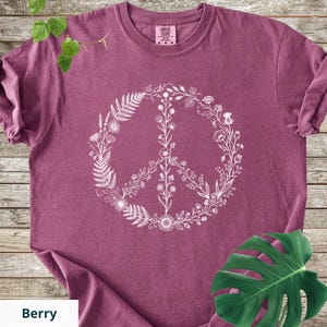 May include: A berry-colored short-sleeve t-shirt featuring a white graphic print of a peace sign composed of various botanical elements, including ferns, wildflowers, and stems. The shirt has a crew neckline and a Comfort Colors brand label.