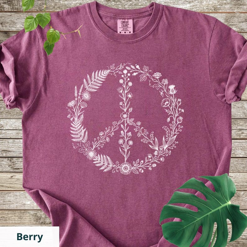 May include: A berry-colored short-sleeve t-shirt featuring a white graphic print of a peace sign composed of various botanical elements, including ferns, wildflowers, and stems. The shirt has a crew neckline and a Comfort Colors brand label.