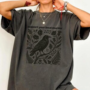 May include: A charcoal gray short-sleeve t-shirt featuring a black graphic print of a crow perched on a branch surrounded by botanical elements. The text "support your local murder" is printed in black above the crow illustration.