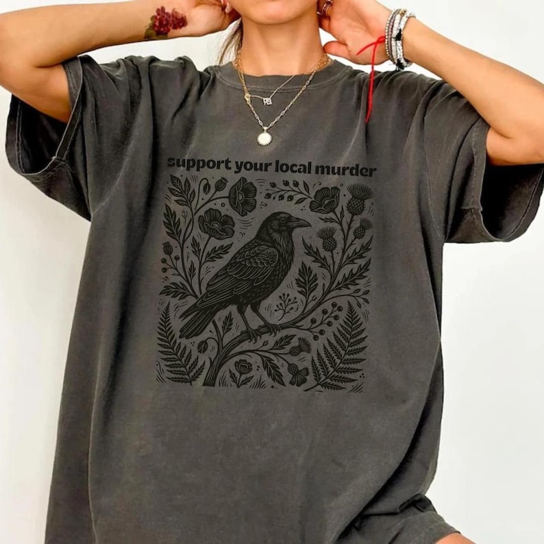 May include: A charcoal gray short-sleeve t-shirt featuring a black graphic print of a crow perched on a branch surrounded by botanical elements. The text "support your local murder" is printed in black above the crow illustration.