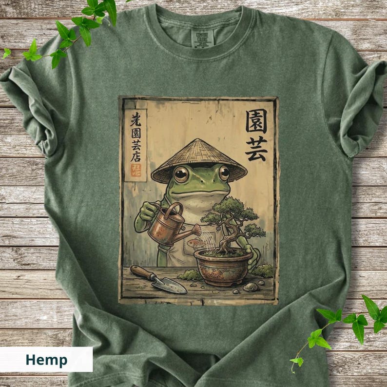 May include: Green t-shirt featuring a frog wearing a conical hat, watering a bonsai tree. The frog is depicted in a Japanese art style. The shirt has the word "Hemp" printed at the bottom. Japanese text is also visible.
