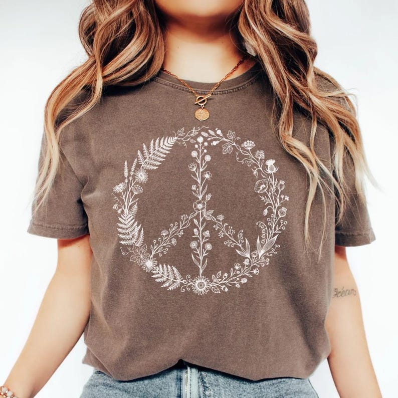 May include: A dark brown short-sleeve t-shirt featuring a white graphic print of a peace sign composed of various botanical elements, including ferns, wildflowers, and stems, centered on the chest area of the garment.
