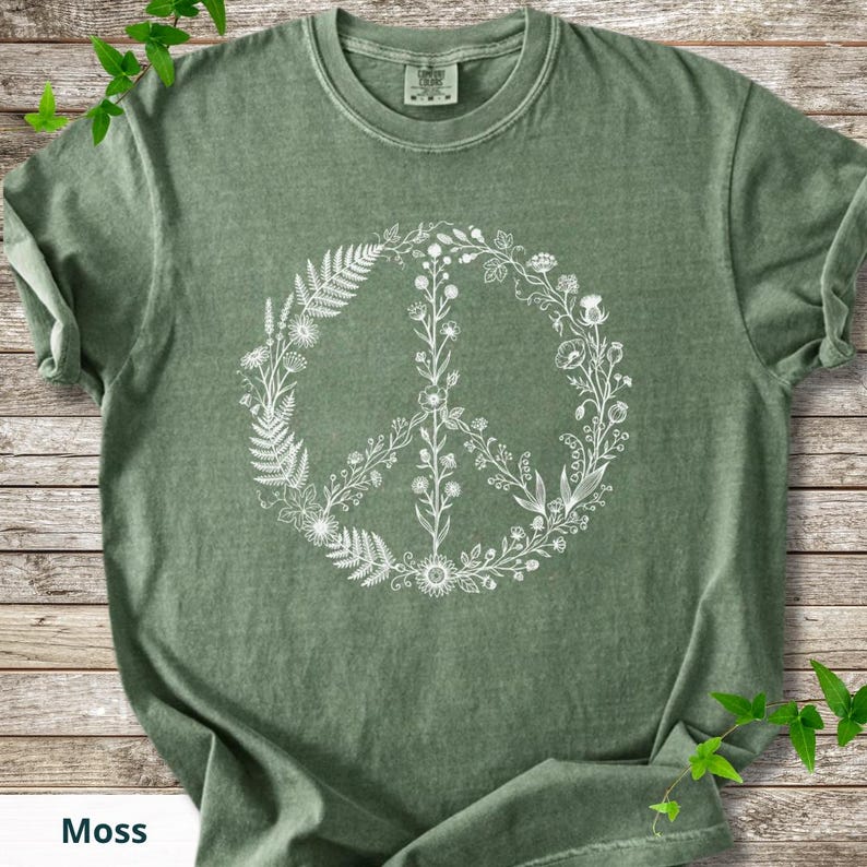 May include: A moss green short-sleeved t-shirt featuring a white graphic print of a peace sign composed of various botanical elements, including ferns, wildflowers, and leafy stems. The shirt has a crew neckline and a visible brand label.