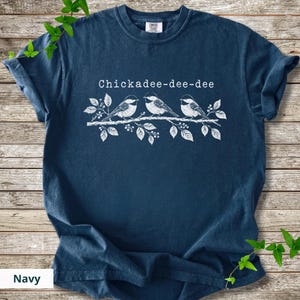 May include: A navy blue short-sleeve t-shirt featuring a white graphic print of three birds perched on a leafy branch. Above the birds, the text reads Chickadee-dee-dee in a white font. The shirt has a crew neckline and a standard fit.