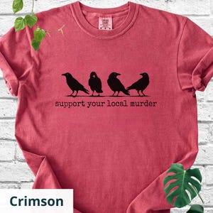 May include: A crimson red short-sleeve t-shirt featuring a black graphic of four crows standing in a row. Below the birds, the text reads "support your local murder" in a typewriter-style font. The shirt has a crew neckline and a visible brand label.