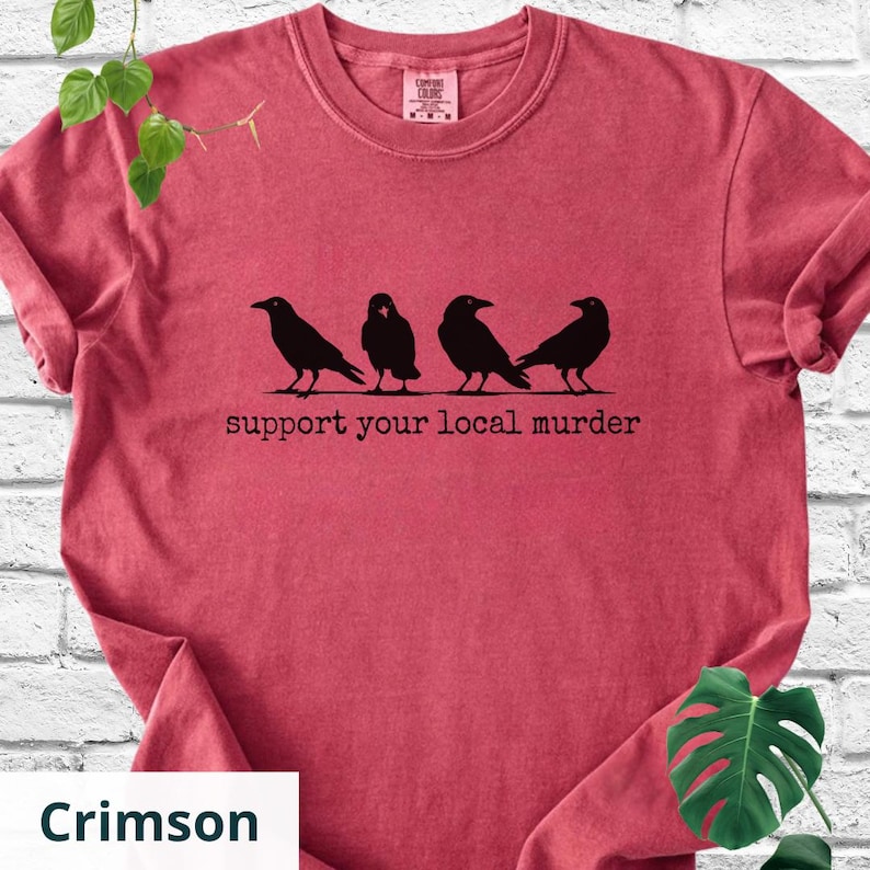 May include: A crimson red short-sleeve t-shirt featuring a black graphic of four crows standing in a row. Below the birds, the text reads "support your local murder" in a typewriter-style font. The shirt has a crew neckline and a visible brand label.