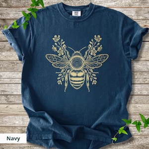 May include: Navy blue t-shirt featuring a detailed golden bee design with floral accents. The bee is centered on the shirt, with wings spread and surrounded by delicate flowers. The shirt is made of soft material.